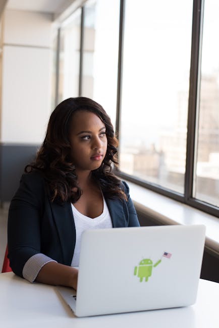 Focused woman using a laptop with Android sticker, sitting by a window in an office.