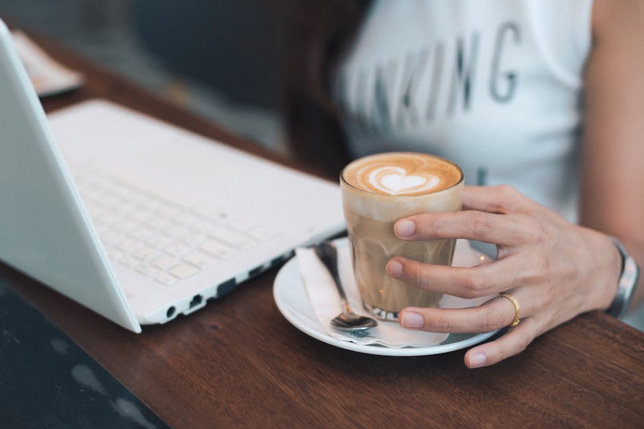 A woman holding a cup of coffee while working on a laptop at a café desk.