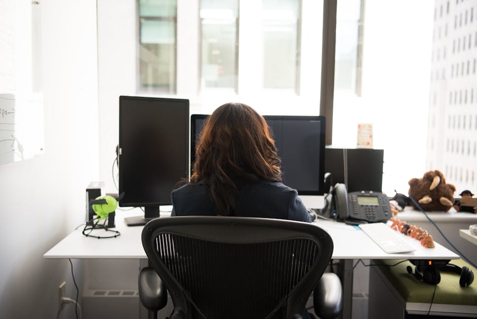 Woman working at a desktop setup in a modern office with technology and city view.
