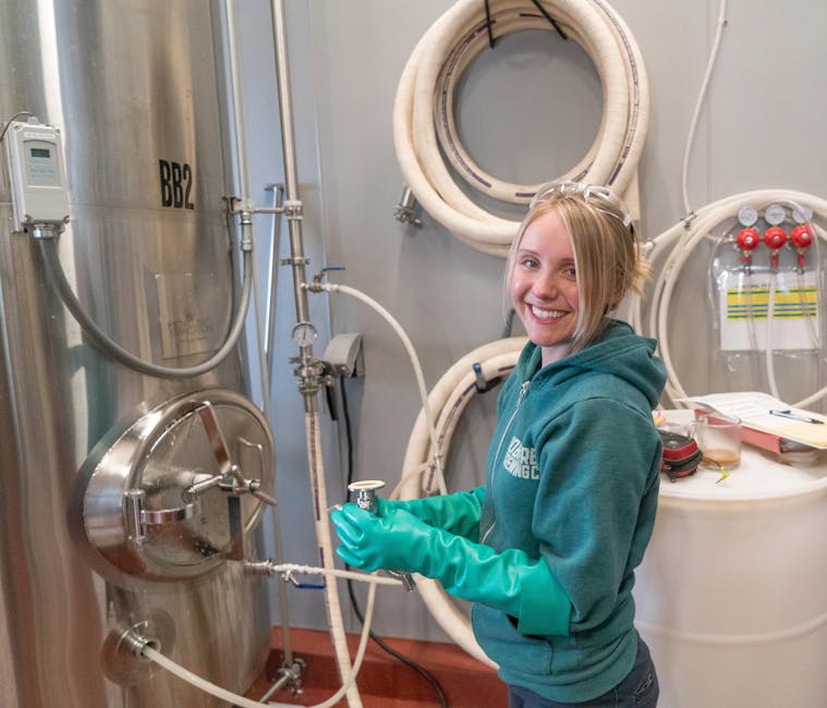 A woman in a green jacket and gloves working in a brewing factory, smiling near stainless steel equipment.